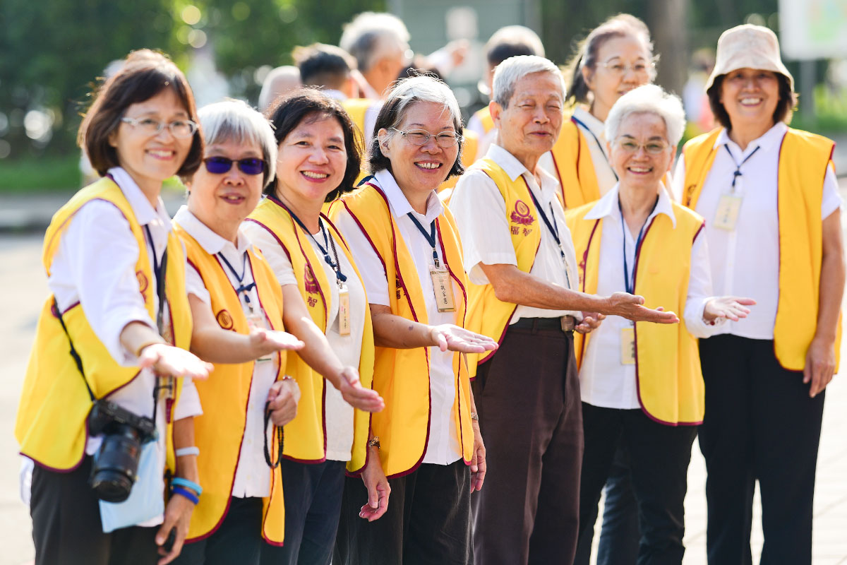 陽光下，身穿黃色義工背心的工作人員面帶微笑，伸手做出邀請報到的姿勢，氣氛熱情且親切